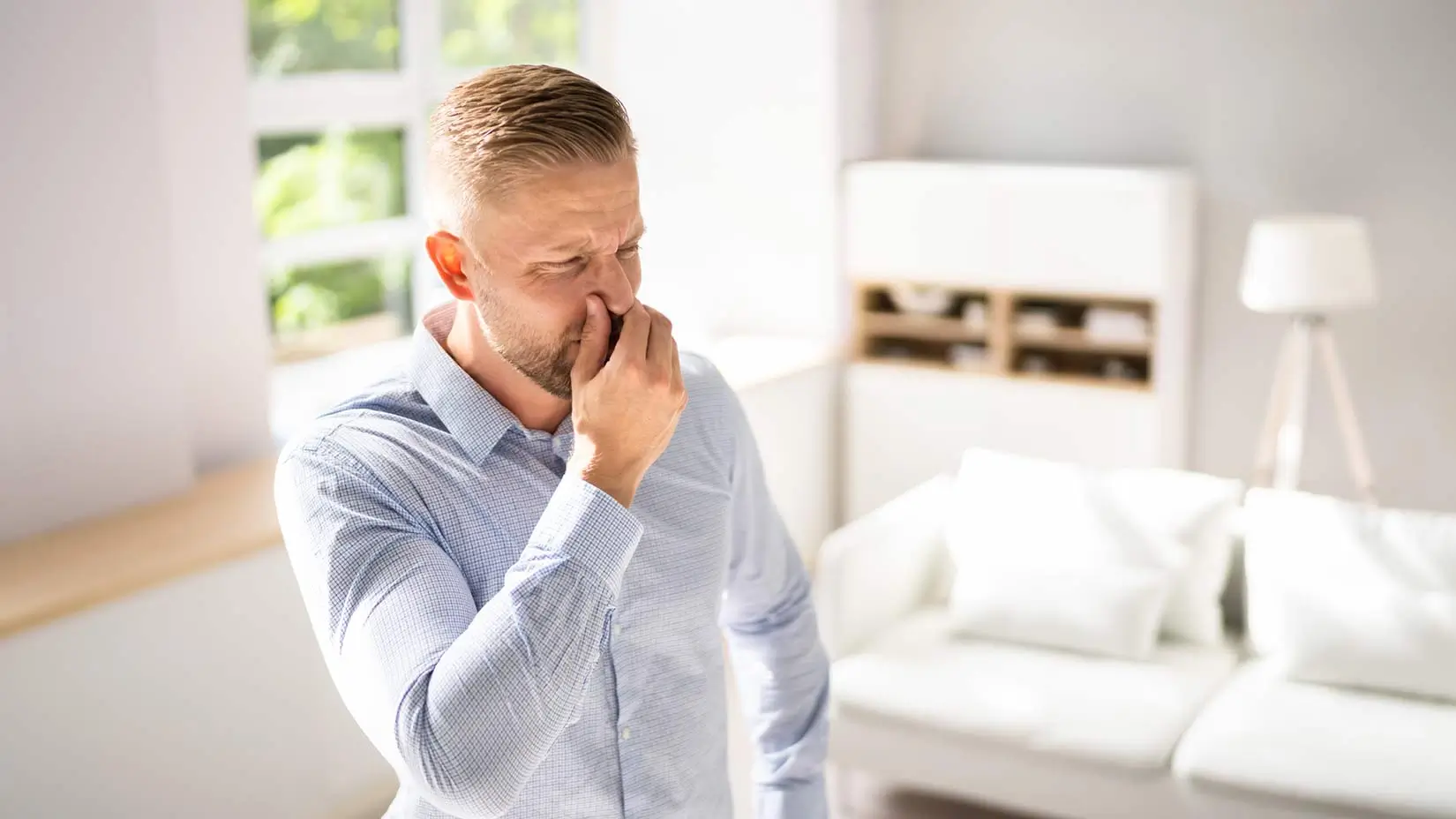 A man stands in his living room plugging his nose and making a disgusted face, presumably because of a sewer gas smell.